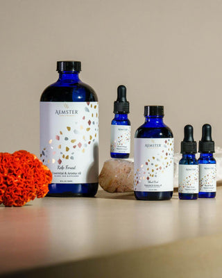 A set of Aemster herbal and aromatherapy bottles arranged on a tabletop, including a large cobalt blue bottle and smaller blue bottles with white labels featuring gold and multicolor confetti. A textured orange coral piece and a pink stone sit beside the products against a warm beige background.